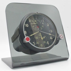 May include: A black aircraft clock with a white face and red accents. The clock has two hands and a small dial with the text "25 CEK" and numbers 5, 10, 15, 20, 25, and 30. The clock is mounted on a clear acrylic stand.