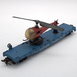 May include: A blue model flatcar train carriage with the text SOUTHERN PACIFIC printed on the side. A red helicopter with a gold-colored cockpit and black rotor blades is mounted on the center of the flatcar, featuring a rotating base.