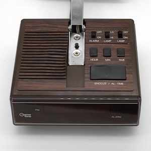 May include: A brown Cosmo Time digital alarm clock with a wood grain finish. The top features a speaker grille, a metal hinge, and black control switches labeled Alarm, Lamp, Hour, Min, Time, and Snooze/Al. Time. The front display shows PM and Alarm.