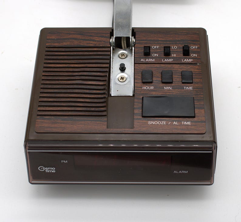 May include: A brown Cosmo Time digital alarm clock with a wood grain finish. The top features a speaker grille, a metal hinge, and black control switches labeled Alarm, Lamp, Hour, Min, Time, and Snooze/Al. Time. The front display shows PM and Alarm.