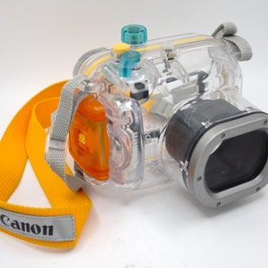 May include: A clear plastic waterproof camera case with a yellow strap and the word "Canon" printed on it. The case has a silver lens cover and a blue and yellow button on the top.