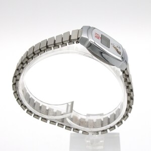 May include: A silver-toned digital wristwatch featuring a metallic expansion band. The watch face displays the red text RCA and a small graphic of a dog listening to a gramophone. The rectangular digital screen is centered within a polished silver case.