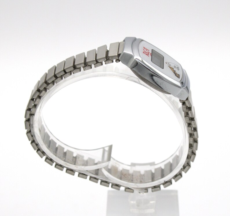 May include: A silver-toned digital wristwatch featuring a metallic expansion band. The watch face displays the red text RCA and a small graphic of a dog listening to a gramophone. The rectangular digital screen is centered within a polished silver case.
