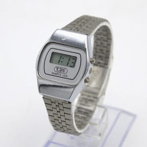 May include: A silver-toned digital watch with a rectangular face displaying the time 1:13. The watch has a silver band and the brand name "TOZAI QUARTZ LCD" is visible on the face. The watch is displayed on a clear stand.