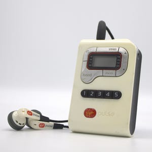 May include: A cream-colored Virgin Pulse portable radio with a black antenna and buttons. The radio has a digital display and comes with white earbuds. The text "Virgin pulse" is printed on the radio.