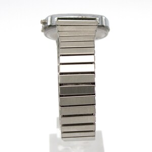 Peut inclure: Une montre-bracelet argent&eacute;e avec un cadran rectangulaire et un bracelet en m&eacute;tal souple. Le bracelet est compos&eacute; de nombreux petits maillons rectangulaires. La montre est pos&eacute;e sur un fond blanc, soulignant son &eacute;clat m&eacute;tallique et son design.