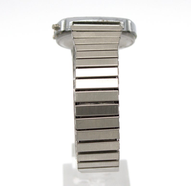 Peut inclure: Une montre-bracelet argent&eacute;e avec un cadran rectangulaire et un bracelet en m&eacute;tal souple. Le bracelet est compos&eacute; de nombreux petits maillons rectangulaires. La montre est pos&eacute;e sur un fond blanc, soulignant son &eacute;clat m&eacute;tallique et son design.