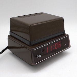 May include: A brown Cosmo Time digital alarm clock with a rectangular plastic housing. The front display features a dark tinted panel with a red LED readout showing 11:06 PM. A black power cord extends from the side of the device.