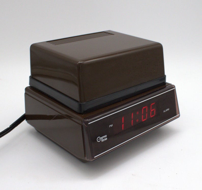 May include: A brown Cosmo Time digital alarm clock with a rectangular plastic housing. The front display features a dark tinted panel with a red LED readout showing 11:06 PM. A black power cord extends from the side of the device.