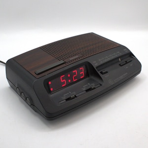 May include: Black and brown digital alarm clock with a red LED display showing 5:23. The clock has a wood-grain top and a speaker grill. It features buttons for snooze and radio settings.