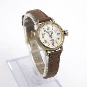May include: A vintage wristwatch with a white face, black numerals, and brown leather band. The watch has a gold-colored bezel and a small crown. The watch face reads "NTANO QUARTZ".