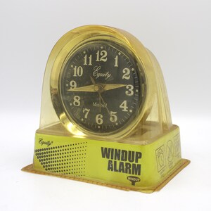 May include: A vintage windup alarm clock with a gold-colored frame and a clear plastic cover. The clock face is black with gold numerals and hands. The text "Equity" and "Windup Alarm" are visible.