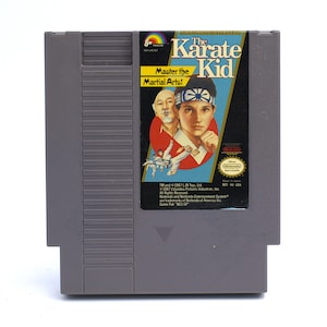 May include: A gray Nintendo Entertainment System (NES) game cartridge for "The Karate Kid." The label features a colorful illustration of the movie's characters and the text "Master the Martial Arts!"