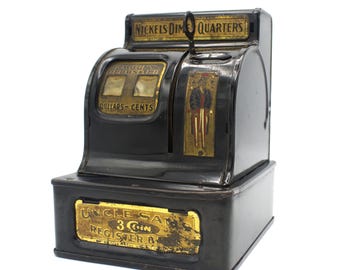 Könnte beinhalten: Schwarze Spielzeug-Kasse aus Metall mit einem goldenen Etikett mit der Aufschrift "Uncle Sam's 3 Coin Register Bank". Die Kasse hat einen Schlitz für Münzen und ein Display mit der Aufschrift "Amount Registered Dollars - Cents".