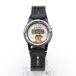 May include: Black digital watch with a white face featuring the brand name "ASAHI" and the time "1:55". The watch face has a cartoon bear illustration. The watch has a black band with the word "SPORT" on it.