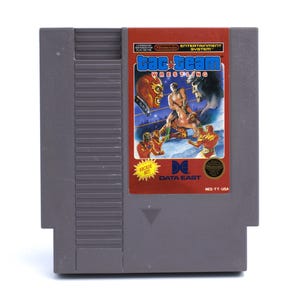 May include: A gray Nintendo Entertainment System (NES) video game cartridge for "Tag Team Wrestling." The label features wrestling figures and the text "Tag Team Wrestling," "Arcade Hit," and "Data East."