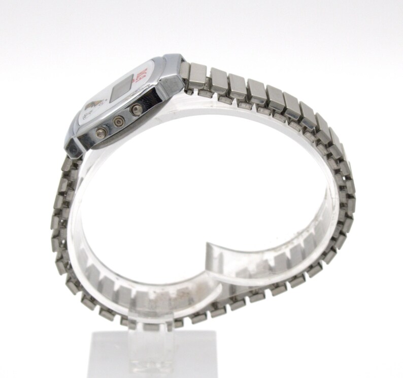 May include: A silver-tone digital wristwatch featuring a rectangular case with a white face, a small digital display, and three side buttons. The watch includes a flexible, segmented metal expansion band with a polished finish.