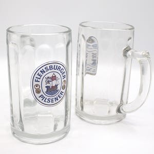 May include: Two clear glass beer mugs. One mug has a blue and gold logo that says "Flensburger Pilsener" with a ship inside the logo.