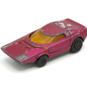 May include: A vintage, die-cast toy car in a faded magenta color. The car features a low-profile design with a yellow windshield and black wheels. The paint shows signs of wear, adding to its retro charm.