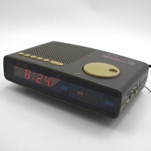 May include: A gray AM/FM digital alarm clock with a red LED display showing 8:24. The clock has a perforated speaker grill, several yellow buttons, and a large tan control dial. The text "am-fm" is visible on the top.