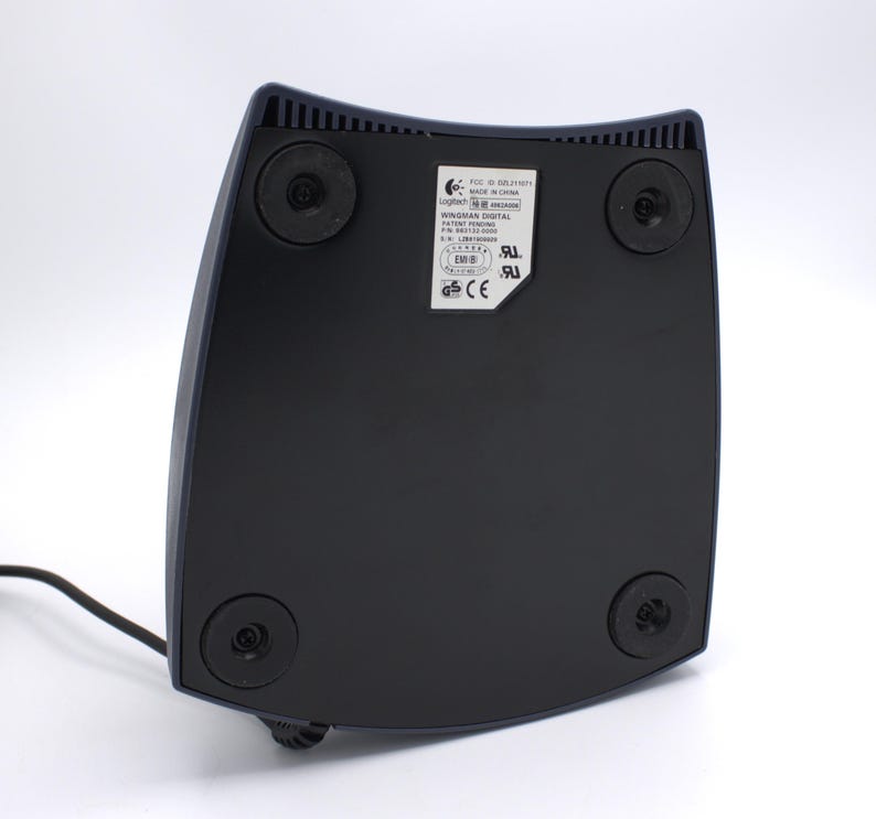 Puede incluir: Base de mando de juegos Logitech WingMan Digital negra y azul marino, vista desde abajo. La base tiene cuatro patas de goma negras y una etiqueta con informaci&oacute;n del producto. Un cable negro se extiende desde la base.