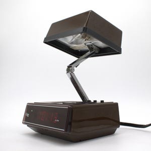May include: A brown plastic alarm clock with a digital red display and an adjustable, hinged metal arm supporting a rectangular brown lamp shade. The base features control buttons and the text "Glow Time" on the front display panel.