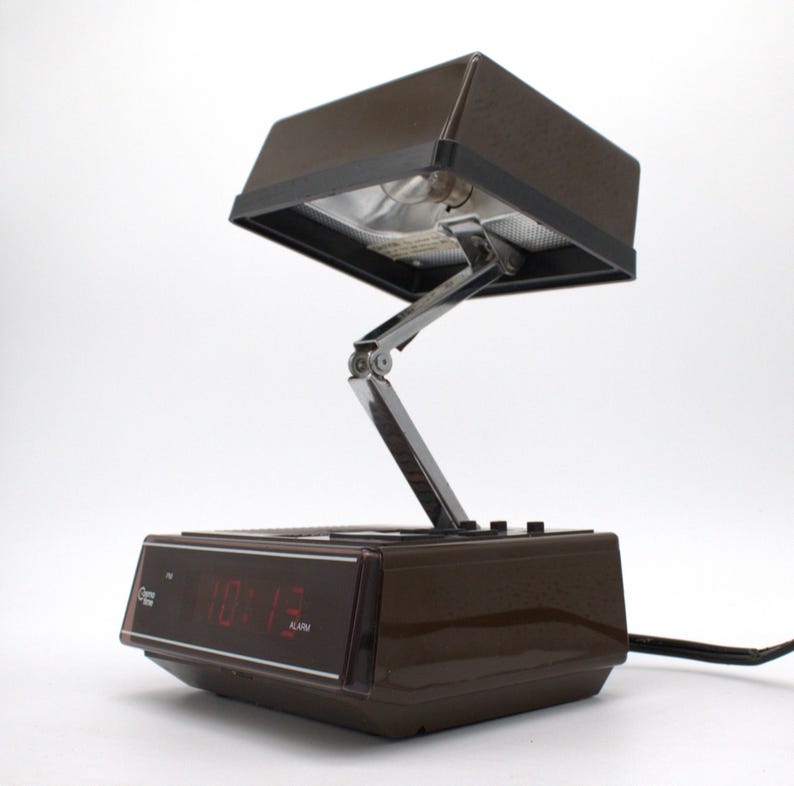 May include: A brown plastic alarm clock with a digital red display and an adjustable, hinged metal arm supporting a rectangular brown lamp shade. The base features control buttons and the text "Glow Time" on the front display panel.