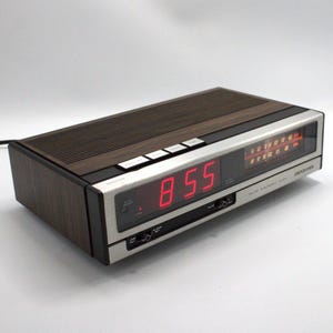 May include: A vintage digital alarm clock with a wood-grain top and sides, a silver face, and a red LED display showing "8:55". The clock has buttons for setting the time and alarm, and a radio dial. The brand name is visible.