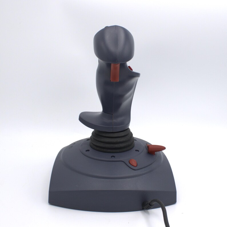 Puede incluir: Un joystick gris oscuro con una base negra y un mango curvo. El joystick tiene un bot&oacute;n rojo y un gatillo rojo. La base tiene un dise&ntilde;o circular con una textura acanalada. Un cable negro se extiende desde la base.