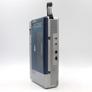 May include: A vintage silver and blue Sony Walkman cassette player. The portable audio device has a black strap, a blue cassette window, and a series of buttons and ports. The word "SONY" is printed on the top.