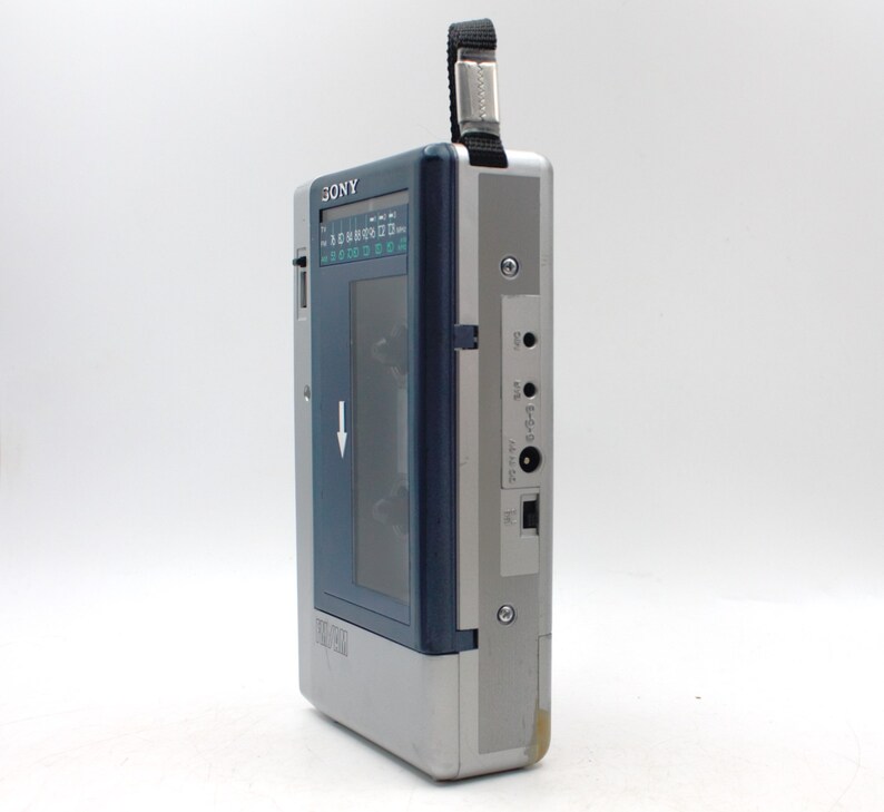 May include: A vintage silver and blue Sony Walkman cassette player. The portable audio device has a black strap, a blue cassette window, and a series of buttons and ports. The word "SONY" is printed on the top.