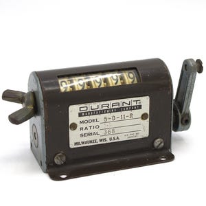 May include: A vintage Durant Manufacturing Company mechanical counter with a dark brown metal casing. The counter displays the numbers 99999. A label on the front reads "Model 5-D-11-R" and "Serial 368". It has a handle and a wing nut.