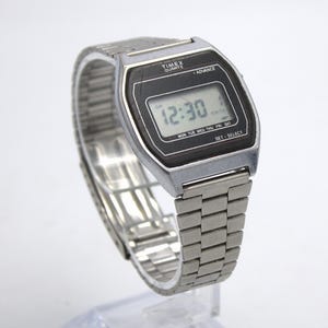 May include: A vintage Timex Quartz digital watch with a silver-tone metal band and black face. The watch displays the time as 12:30 AM and the day of the week. The watch has the text "Timex Quartz Advance" on the face.