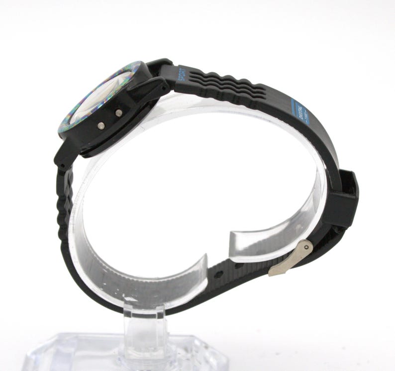 May include: Black sports watch with a textured black band and a multi-colored face. The watch has a digital display and a buckle closure. The band has the word "SPORT" printed on it.