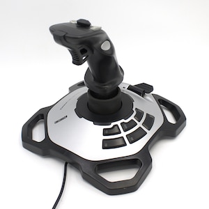 May include: A black and silver joystick controller with multiple buttons and a central control stick. The base has a unique shape with integrated handles. The controller is designed for gaming and has a sleek, modern appearance.