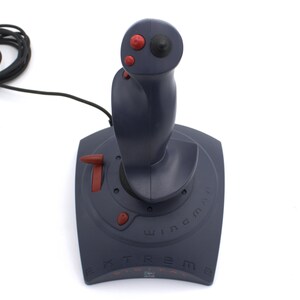 Puede incluir: Un joystick gris oscuro con botones rojos y una parte superior negra. La base del joystick tiene las palabras "Extreme Digital" y "Wingman" impresas. Un cable negro se extiende desde la parte superior del joystick.