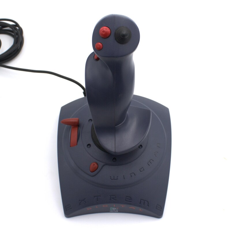 Puede incluir: Un joystick gris oscuro con botones rojos y una parte superior negra. La base del joystick tiene las palabras "Extreme Digital" y "Wingman" impresas. Un cable negro se extiende desde la parte superior del joystick.