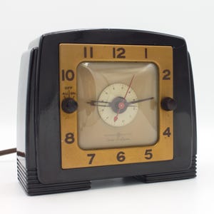 May include: A black and gold vintage alarm clock with a square face and a white dial. The clock has a black plastic case and a gold-colored bezel. The clock face has black numbers and hands, and a small red second hand. The clock has a knob on the right side for setting the alarm.
