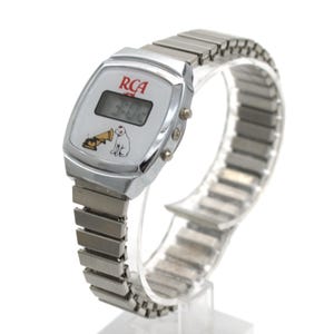 May include: A silver-tone digital wristwatch with a metallic expansion band. The white watch face features the red RCA logo, a digital time display, and a graphic illustration of the Nipper dog listening to a gramophone horn.
