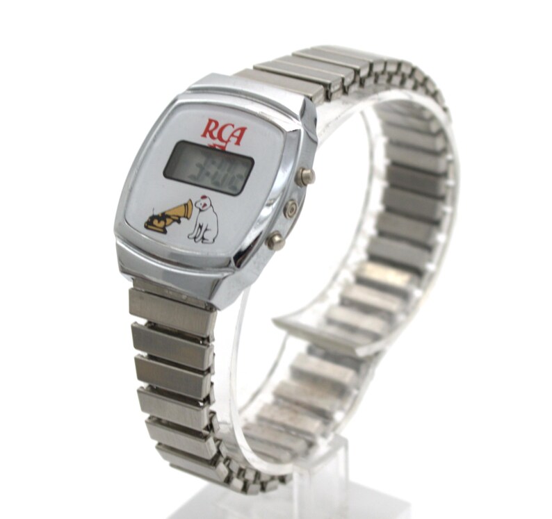May include: A silver-tone digital wristwatch with a metallic expansion band. The white watch face features the red RCA logo, a digital time display, and a graphic illustration of the Nipper dog listening to a gramophone horn.