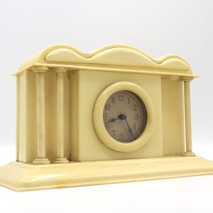 May include: A cream-colored, decorative clock with a Roman numeral face and a design resembling a miniature building with columns.