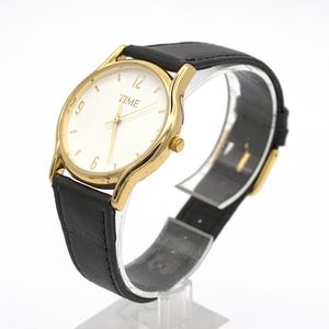 May include: Gold-tone wristwatch with a white face and black leather band. The watch face displays the word "TIME" and simple hour markers. The watch is displayed on a clear stand.