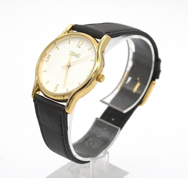 May include: Gold-tone wristwatch with a white face and black leather band. The watch face displays the word "TIME" and simple hour markers. The watch is displayed on a clear stand.