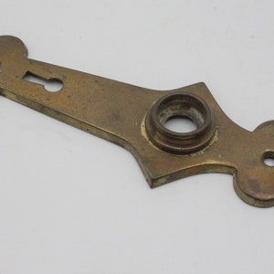 May include: A vintage brass door plate with a keyhole and a circular opening. The plate has a decorative shape with rounded edges and a slightly tarnished finish, suggesting age and use. It is likely a component of antique hardware.