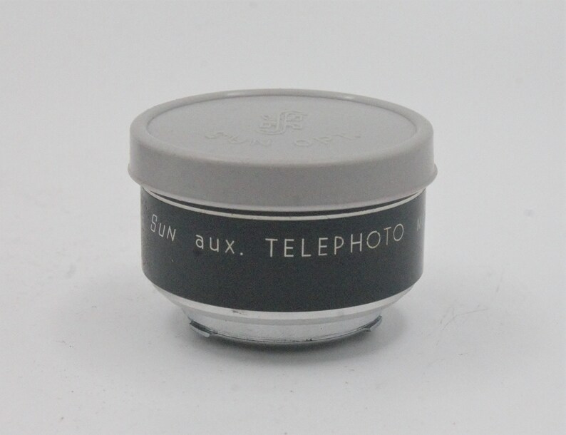 May include: A black and silver telephoto lens attachment for a camera. The lens has a gray plastic cap with the text "Sun aux. Telephoto" printed on the side.