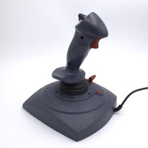 Puede incluir: Un joystick gris con un bot&oacute;n y detalles en rojo. El joystick tiene un cable negro y una base con n&uacute;meros y letras. El joystick est&aacute; dise&ntilde;ado para juegos.