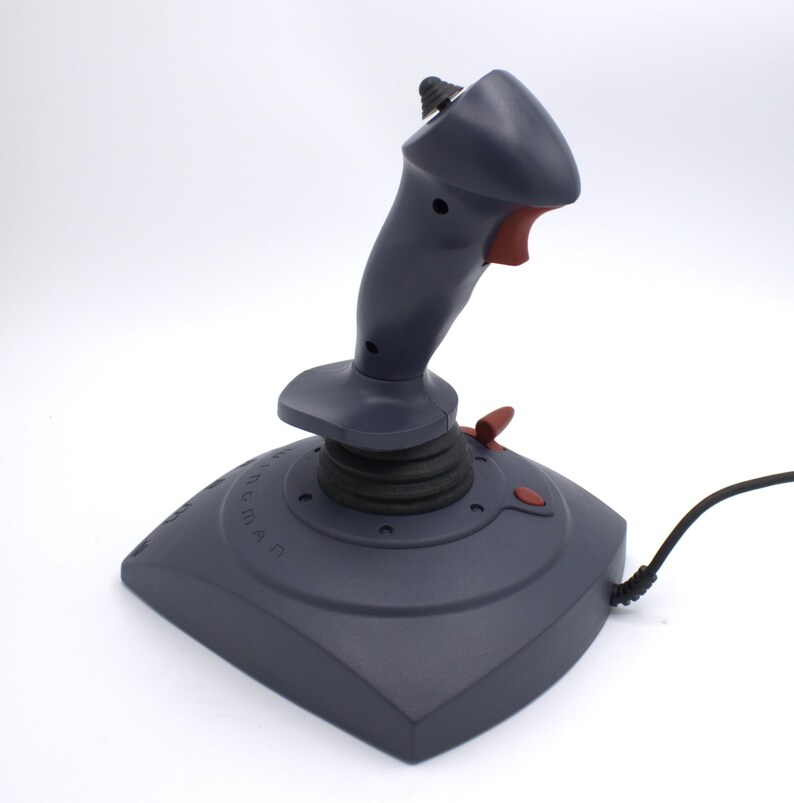 Puede incluir: Un joystick gris con un bot&oacute;n y detalles en rojo. El joystick tiene un cable negro y una base con n&uacute;meros y letras. El joystick est&aacute; dise&ntilde;ado para juegos.