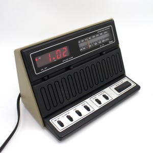 May include: A vintage digital alarm clock with a beige and black color scheme. The clock displays the time in red digits and has an AM/FM radio. It features buttons for time and alarm settings, and a battery backup.