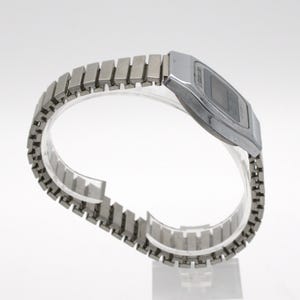 May include: A silver-toned digital wristwatch featuring a rectangular case and a flexible, expandable metal link bracelet. The watch face has a dark display screen with visible text reading SKELLIN, set within a polished metallic frame.