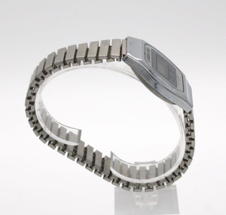 May include: A silver-toned digital wristwatch featuring a rectangular case and a flexible, expandable metal link bracelet. The watch face has a dark display screen with visible text reading SKELLIN, set within a polished metallic frame.